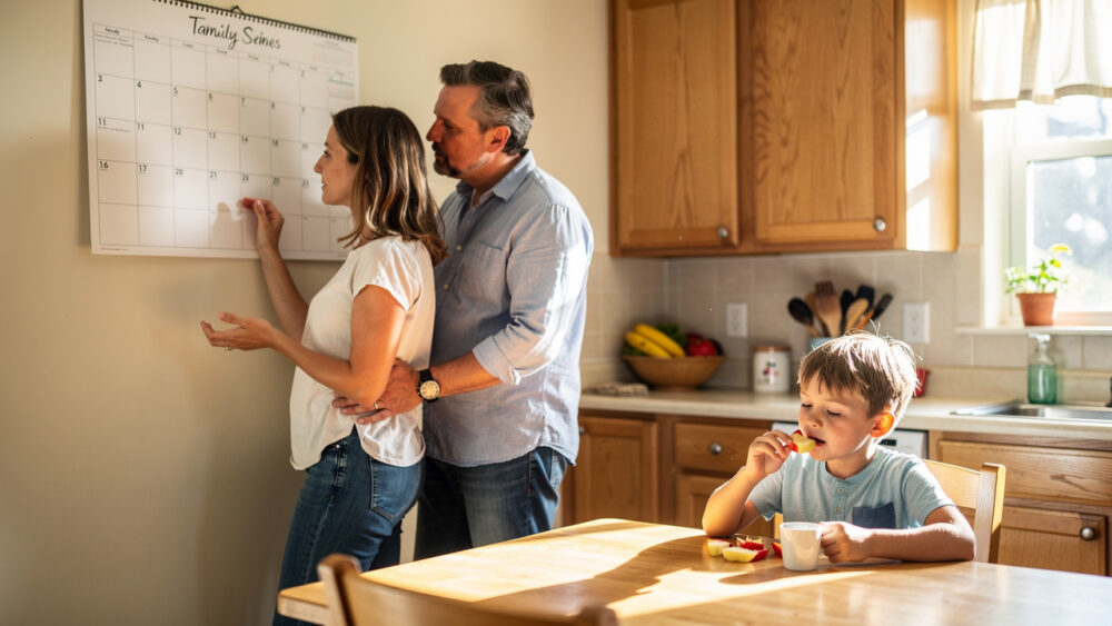 Un père et une mère consultent un calendrier familial vierge dans une cuisine lumineuse, tandis que leur jeune garçon prend son goûter dans une ambiance chaleureuse.